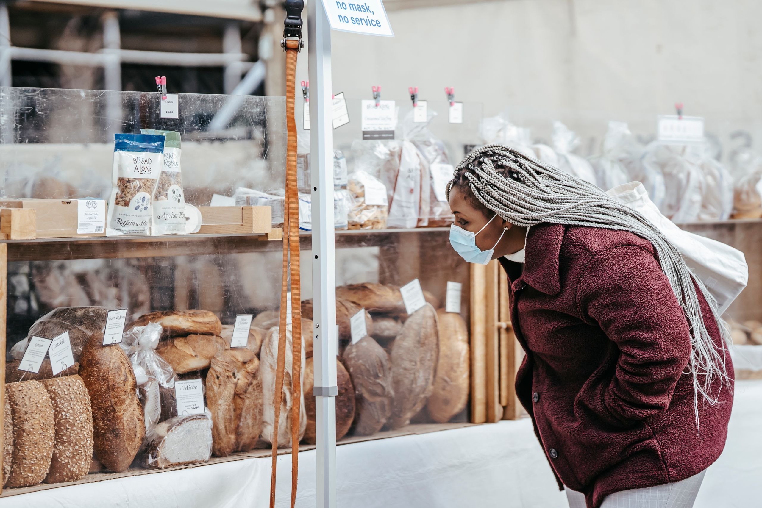 Why I Keep Looking for Local Bread - Side view of adult black lady wearing protective mask and warm coat selecting baking goods while standing in street market in daytime
