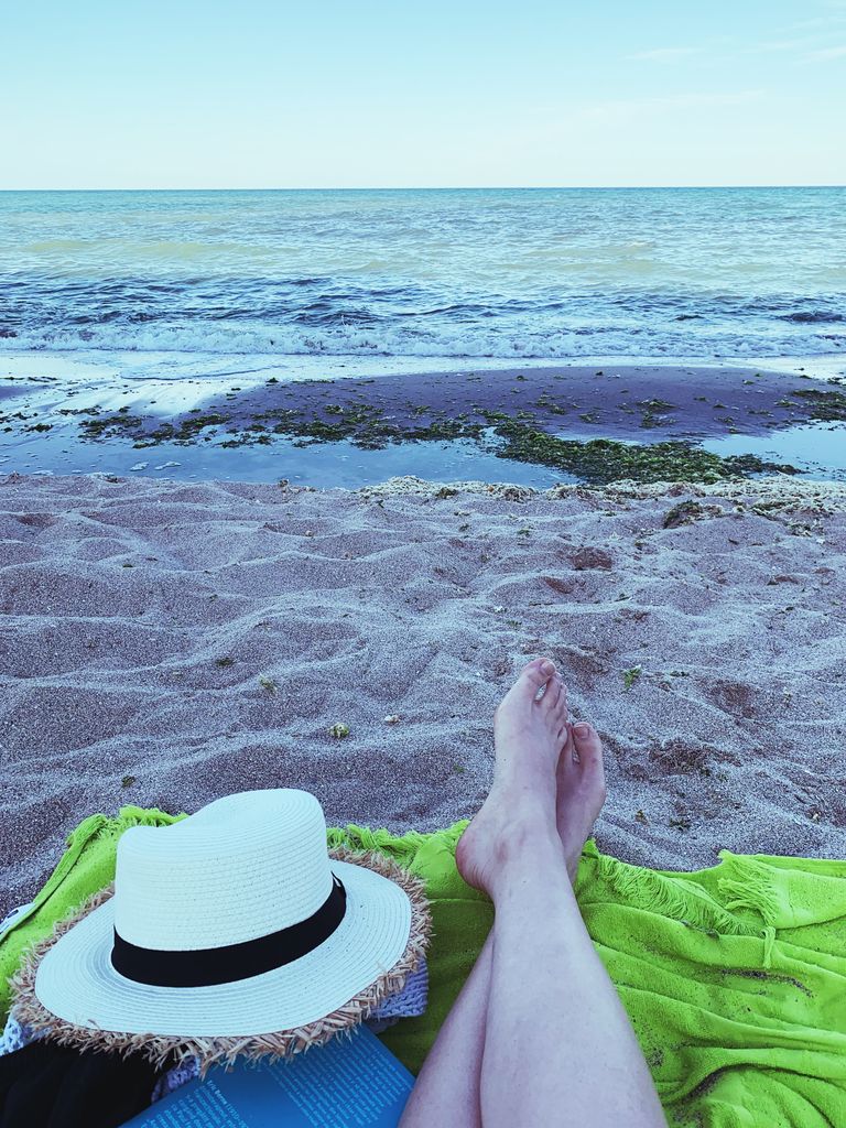 Why I Still Believe in Summer Even When It Is Complicated - Crop barefoot person with straw hat lying on green plaid on sandy shore near blue waving sea