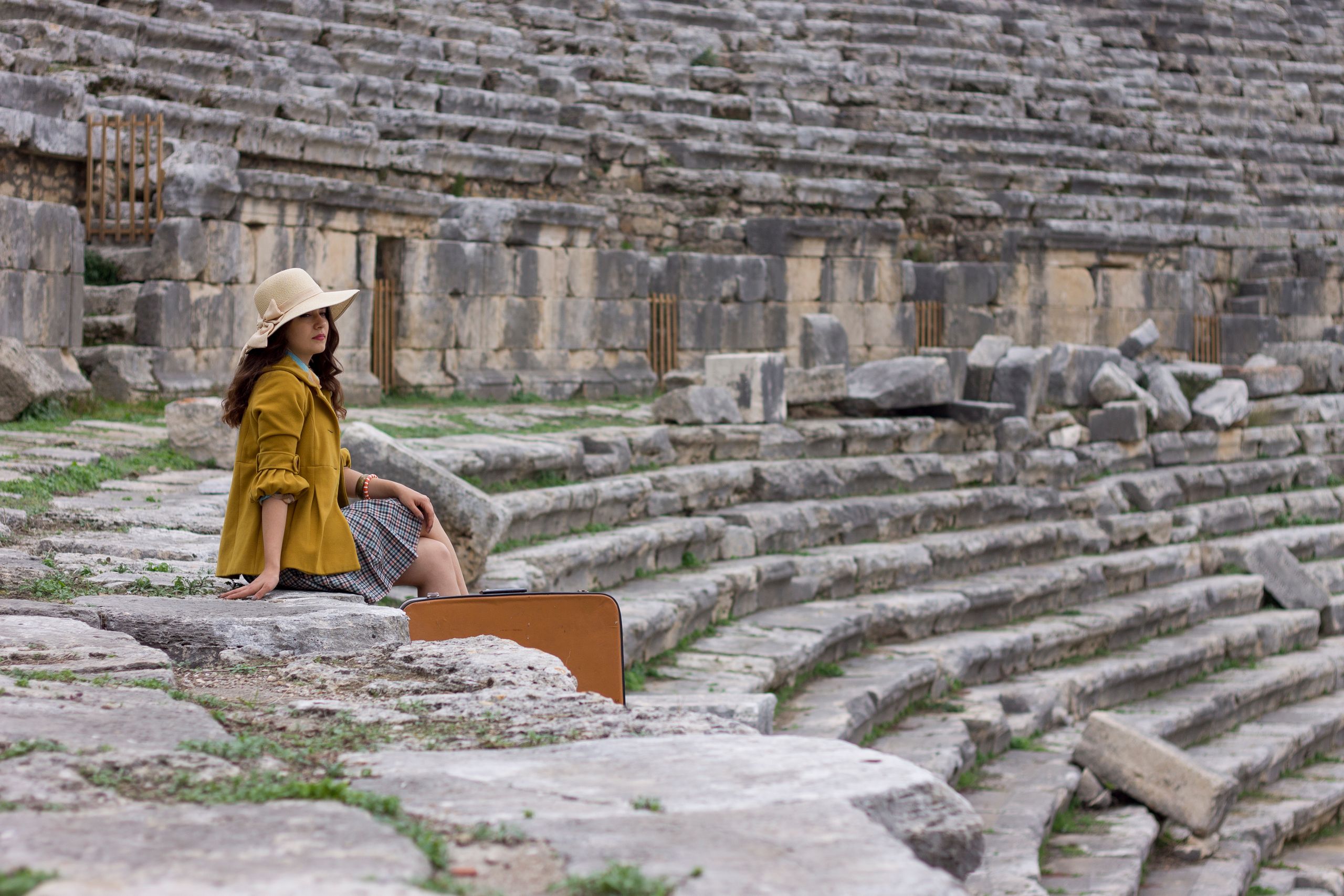 Why I Travel for Culture, Not Escape | DG Speaks - A woman in a hat and yellow jacket sitting in an ancient Turkish amphitheater.