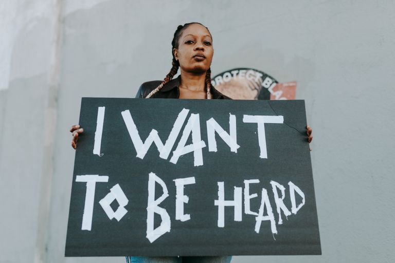Why I Want Rooms That Let Women Be Loud - A young woman holds a protest sign outdoors, advocating for being heard.