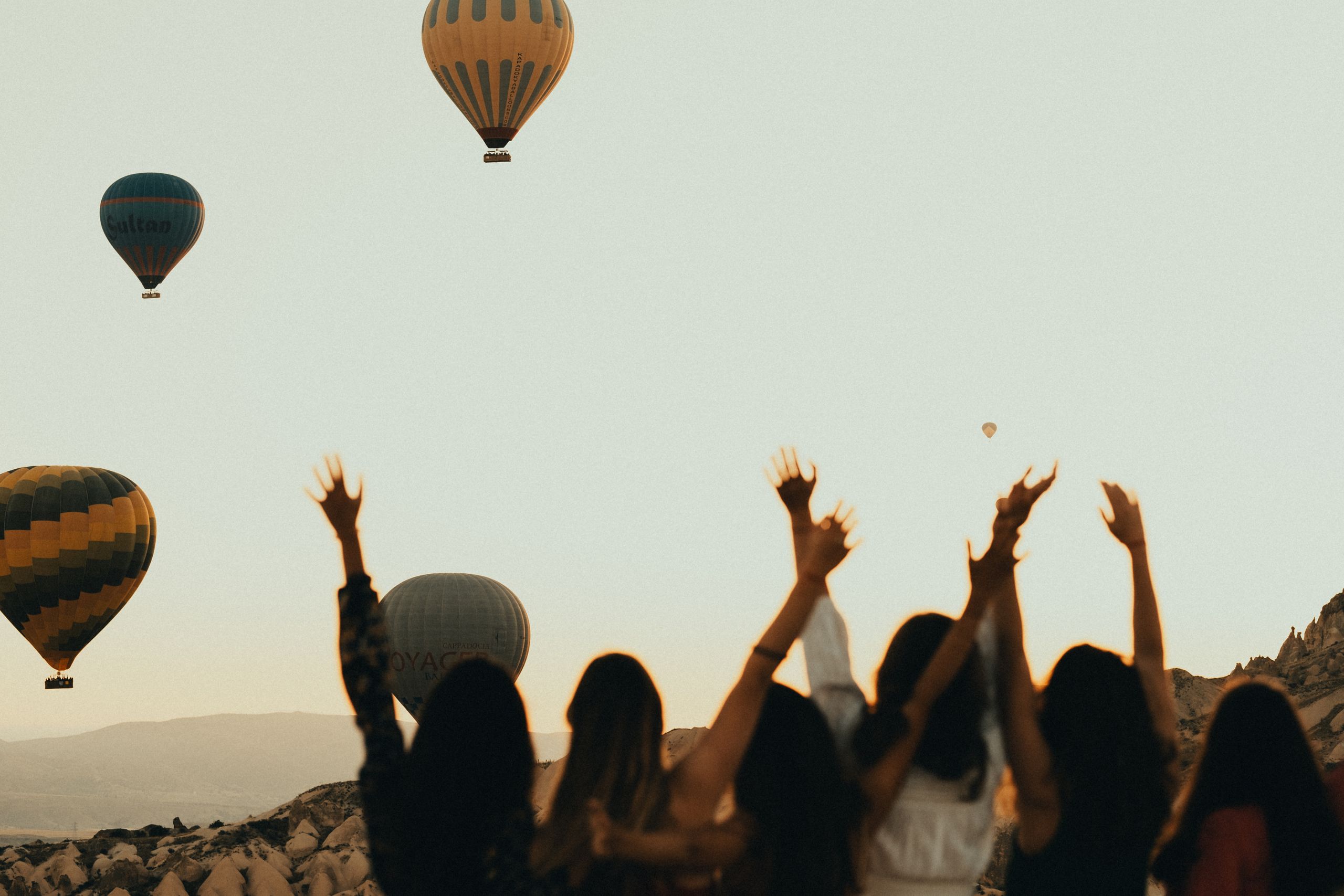 Women Who Make Travel Feel Possible - Silhouettes of women watching hot air balloons at sunset, arms raised in excitement.