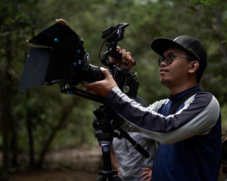 A man in a cap operates a professional camera rig in a forest environment, filming outdoors.